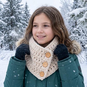 May include: A cream-colored chunky knit cowl scarf featuring three round wooden buttons arranged vertically along the front edge. The scarf is worn over a dark green winter jacket with a fur-trimmed hood and black knit gloves in a snowy setting.