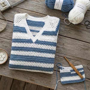 May include: A V-neck sweater featuring horizontal stripes in white and dusty blue yarn. The garment has a textured crochet stitch pattern. Included are two skeins of yarn, a wooden crochet hook, and a small swatch of matching striped fabric.