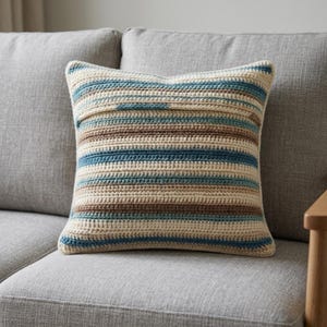 Könnte beinhalten: Ein quadratisches Häkelkissen mit horizontalen Streifen in den Farben Creme, Hellbraun und Blaugrün. Das Textil weist ein sichtbares, strukturiertes Garnstichmuster auf, das ein durchgehendes, sich wiederholendes Streifendesign auf der Vorderseite erzeugt.