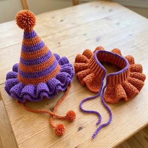 May include: A crocheted cone-shaped hat with orange and purple horizontal stripes, a ruffled purple brim, and an orange pom-pom on top. Beside it is a matching crocheted neck ruff with orange and purple sections and long purple ties.