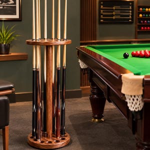 Op de afbeelding: Een ronde, vrijstaande houten biljartkeurek met een centrale kolom en een voet met meerdere uitsparingen. Het rek bevat diverse lange, slanke biljartkeus met lichtgekleurde houten shafts en donkere, gedecoreerde onderstukken in een cirkel.