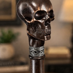 May include: A dark brown wooden walking stick featuring a carved skull handle. The skull has visible wood grain and deep-set eye sockets. Below the skull is a silver-colored metal collar with an intricate woven knot pattern and etched details.