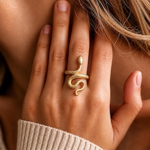 Peut inclure: Une bague dorée en forme de serpent enroulé avec une surface texturée rappelant des écailles. Le design du serpent s'enroule autour du doigt, présentant une tête définie sur le dessus et une queue courbée en bas, formant une silhouette sinueuse.