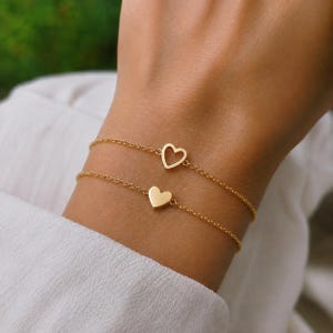 May include: Two delicate gold-toned chain bracelets, each featuring a small heart charm. One bracelet displays an open-frame heart outline, while the other features a solid, smooth heart-shaped charm. Both bracelets are crafted with fine links.