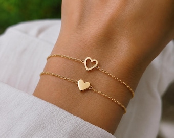 May include: Two delicate gold-toned chain bracelets, each featuring a small heart charm. One bracelet displays an open-frame heart outline, while the other features a solid, smooth heart-shaped charm. Both bracelets are crafted with fine links.