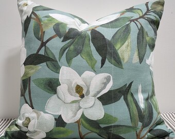 Könnte beinhalten: Ein quadratisches Zierkissen mit einem botanischen Druck aus großen weißen Magnolienblüten mit gelben Zentren und dunkelgrünen Blättern auf hellblauem Grund. Der Stoff weist eine sichtbare Webstruktur auf der gesamten Oberfläche auf.
