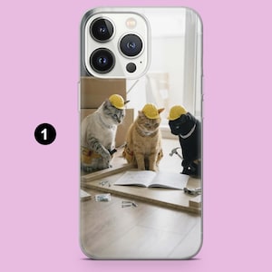 May include: A clear smartphone case featuring a graphic of three cats wearing yellow construction hard hats and tool belts. The cats are positioned around a wooden board and an open instruction manual, with one cat holding a small metal hammer.