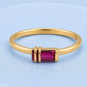 May include: A gold ring featuring a single rectangular baguette-cut magenta gemstone set horizontally. The band is smooth and polished, with two thin vertical gold bars accenting the side of the gemstone setting, creating a clean and simple design.