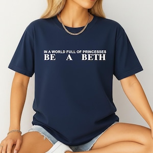 Könnte beinhalten: Ein dunkelblaues Kurzarm-T-Shirt mit weißem Textaufdruck auf der Brust, der lautet: IN A WORLD FULL OF PRINCESSES BE A BETH. Das T-Shirt hat einen Rundhalsausschnitt und eine Standardpassform, was für einen schlichten Look sorgt.