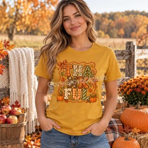 Può includere: Una t-shirt a maniche corte color giallo senape con la scritta THANKFUL in grandi lettere stampate. Il design è circondato da illustrazioni a tema autunnale, tra cui zucche arancioni, foglie autunnali e piccoli fiori.