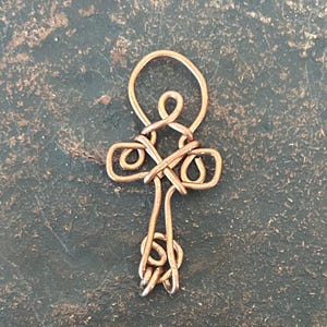 Può includere: Un ciondolo in filo di rame a forma di ankh, caratterizzato da una parte superiore ad anello, una sezione centrale a croce con dettagli in filo intrecciato e una base decorativa a spirale. Il metallo ha un caldo tono ramato.