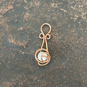 Può includere: Un ciondolo in filo di rame caratterizzato da un occhiello circolare in alto, due piccoli riccioli simmetrici sottostanti e una base a spirale più ampia che racchiude una singola perlina bianca traslucida, rotonda e piccola.