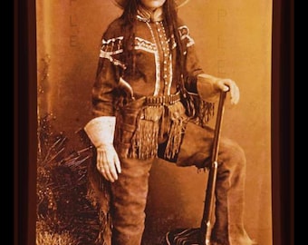 May include: A sepia-toned photograph of a woman wearing a wide-brimmed hat, a long-sleeved shirt with fringe, and pants. She is holding a rifle and standing on a patterned rug.