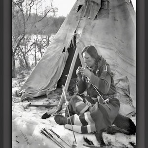 May include: A black and white photograph of a person sitting in front of a teepee, working on a project. The person is wearing a traditional outfit and is surrounded by tools and supplies. The image is taken in a snowy setting.