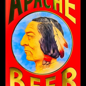 May include: A vintage-style metal sign advertising Apache Beer. The sign features a red background with gold lettering and a circular image of a Native American man with a blue sky background. The text on the sign reads "Wild Enough To Shoot At Apache Beer" and "Made in USA".
