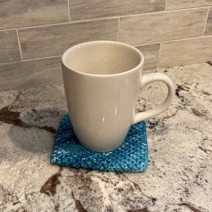 Könnte beinhalten: Eine weiße Keramiktasse mit Henkel auf einem blauen gehäkelten Untersetzer.