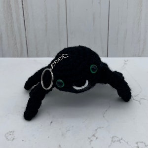 May include: A black crocheted spider keyring with green eyes and a smiling mouth. The spider has four legs and a silver keyring attached to its body. The background is a white and grey surface.