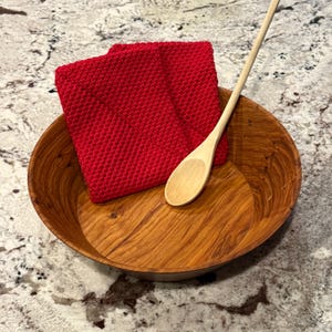 May include: A round wooden bowl with a natural grain pattern, containing a bright red square knitted potholder and a long-handled light-colored wooden spoon resting inside the bowl against the side of the red potholder.