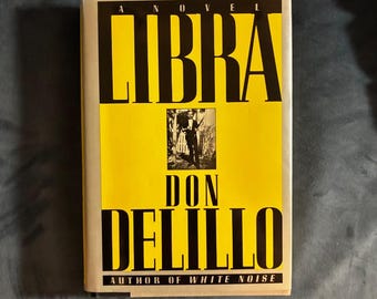 May include: A hardcover book titled Libra by Don DeLillo. The cover features a bright yellow background with bold black text. A small black and white photograph of a person is centered between the title and the author's name.