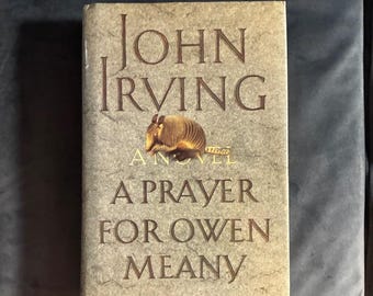May include: A hardcover book titled A Prayer for Owen Meany by John Irving. The cover is a textured beige with brown lettering and features a central illustration of a brown armadillo. The title and author name are printed in large font.