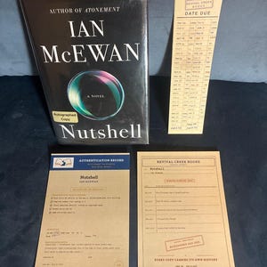 May include: A hardcover book titled Nutshell by Ian McEwan with a black dust jacket featuring a circular graphic, accompanied by a vintage-style library date due card, an authentication record document, and a Revival Creek Books provenance card.