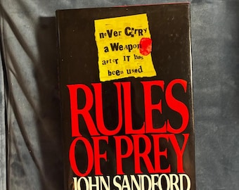 Könnte beinhalten: Ein gebundenes Buch mit dem Titel Rules of Prey von John Sandford mit schwarzem Einband. Oben mittig befindet sich eine gelbe Notizzettel-Grafik mit dem Text: neVer CaRry a WeApon afteR IT has been used, inklusive eines roten Kreises.
