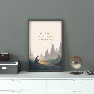 May include: A framed vertical wall art print featuring a beige background with the text "WORDS OUTLAST EMPIRES" centered above a dark silhouette of a person sitting on a rock in a desolate, ruined landscape with jagged, crumbling structures.