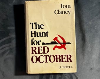 May include: A hardback book titled The Hunt for Red October by Tom Clancy with a cream-coloured dust jacket. The cover features dark brown text, a black silhouette of a submarine, and a red hammer and sickle symbol centred on the front.