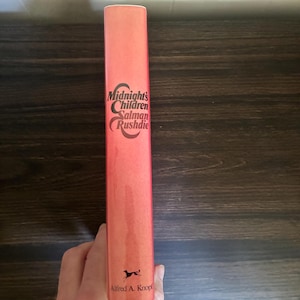 K&ouml;nnte beinhalten: Der Buchr&uuml;cken eines gebundenen Buches mit korallenfarbenem Hintergrund. Der Text Midnight's Children von Salman Rushdie ist in schwarzer Schrift gedruckt. Unten auf dem Buchr&uuml;cken befindet sich eine kleine schwarze Grafik eines Hundes und der Text Alfred A. Knopf.