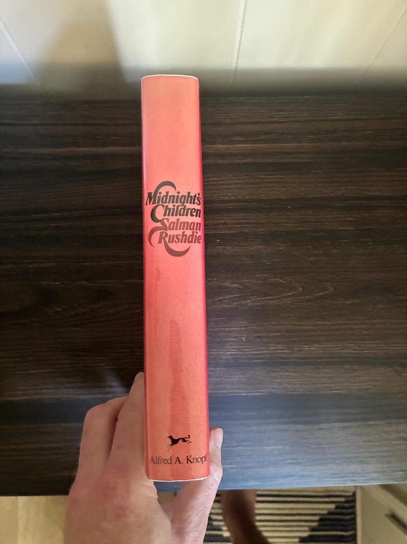K&ouml;nnte beinhalten: Der Buchr&uuml;cken eines gebundenen Buches mit korallenfarbenem Hintergrund. Der Text Midnight's Children von Salman Rushdie ist in schwarzer Schrift gedruckt. Unten auf dem Buchr&uuml;cken befindet sich eine kleine schwarze Grafik eines Hundes und der Text Alfred A. Knopf.