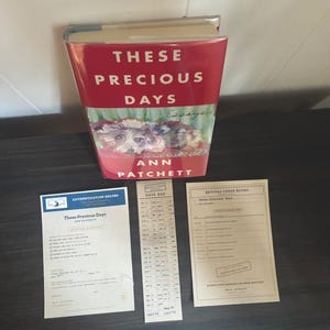 May include: A hardcover book titled These Precious Days by Ann Patchett with a red cover featuring a dog illustration, accompanied by a blue and white authentication record, a vertical date due card, and a beige Revival Creek Books information sheet.