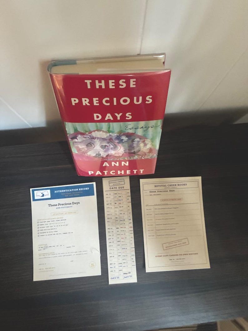 May include: A hardcover book titled These Precious Days by Ann Patchett with a red cover featuring a dog illustration, accompanied by a blue and white authentication record, a vertical date due card, and a beige Revival Creek Books information sheet.