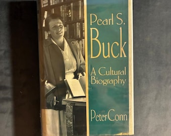Op de afbeelding: Een hardcover boek getiteld Pearl S. Buck: A Cultural Biography door Peter Conn. De stofomslag bevat een zwart-witfoto van Pearl S. Buck voor een boekenplank, met een verticaal blauwgroen paneel met gouden tekst.