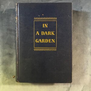 Puede incluir: Un libro de tapa dura azul oscuro con letras doradas en la portada que dice IN A DARK GARDEN. El título está encerrado dentro de un borde rectangular dorado fino con patrones de filigrana decorativos en los bordes superior e inferior.