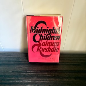 K&ouml;nnte beinhalten: Ein gebundenes Buch mit dem Titel Midnight's Children von Salman Rushdie. Der Buchumschlag ist leuchtend rot mit einer fetten, schwarzen, stilisierten Typografie, die den Titel und den Namen des Autors zeigt. Unten steht der Text A NOVEL.