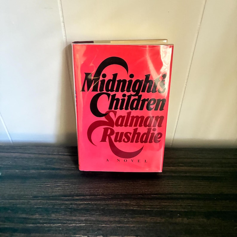 K&ouml;nnte beinhalten: Ein gebundenes Buch mit dem Titel Midnight's Children von Salman Rushdie. Der Buchumschlag ist leuchtend rot mit einer fetten, schwarzen, stilisierten Typografie, die den Titel und den Namen des Autors zeigt. Unten steht der Text A NOVEL.