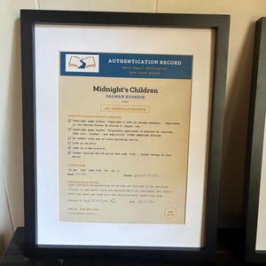 K&ouml;nnte beinhalten: Ein gerahmtes Echtheitszertifikat f&uuml;r die erste amerikanische Ausgabe von Midnight's Children von Salman Rushdie aus dem Jahr 1981. Das Dokument hat eine blaue Kopfzeile, einen beigen Hintergrund und schwarzen Text zu Identifikationsmerkmalen.