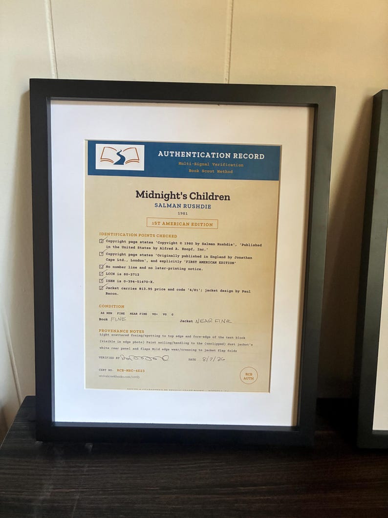 K&ouml;nnte beinhalten: Ein gerahmtes Echtheitszertifikat f&uuml;r die erste amerikanische Ausgabe von Midnight's Children von Salman Rushdie aus dem Jahr 1981. Das Dokument hat eine blaue Kopfzeile, einen beigen Hintergrund und schwarzen Text zu Identifikationsmerkmalen.