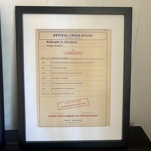 K&ouml;nnte beinhalten: Ein gerahmtes Dokument von Revival Creek Books, das die Geschichte des Buches Midnight's Children von Salman Rushdie detailliert. Das beige Papier zeigt eine Zeitleiste von 1981 bis 2026, einen roten Stempel mit Accessioned Aug 2026 und schwarzen Text.