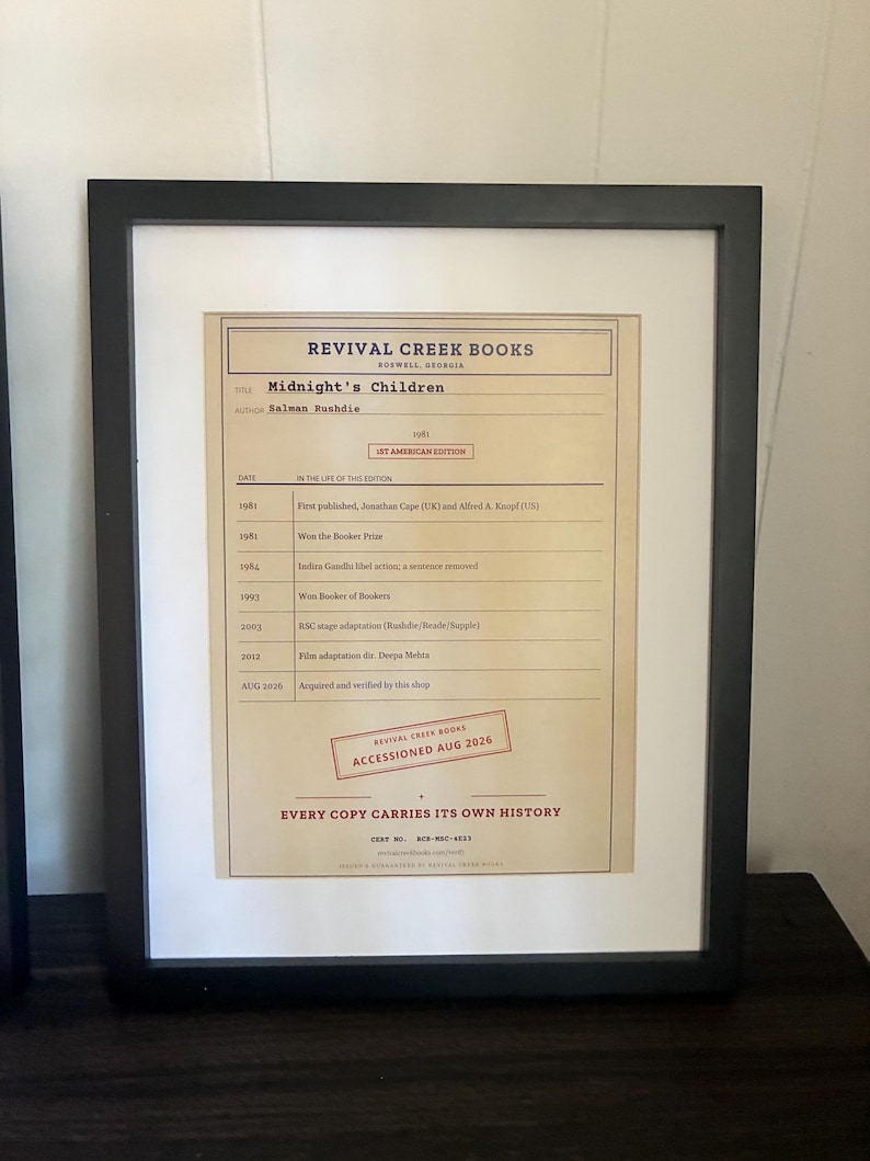 K&ouml;nnte beinhalten: Ein gerahmtes Dokument von Revival Creek Books, das die Geschichte des Buches Midnight's Children von Salman Rushdie detailliert. Das beige Papier zeigt eine Zeitleiste von 1981 bis 2026, einen roten Stempel mit Accessioned Aug 2026 und schwarzen Text.