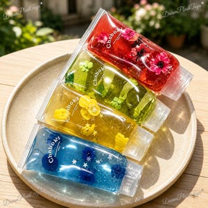 Può includere: Quattro tubetti trasparenti riempiti con gel colorato ed elementi floreali incorporati. I tubetti sono rossi, verdi, gialli e blu, ognuno etichettato con il testo Chnboki. Il gel contiene piccoli glitter a forma di stella e petali di fiori.