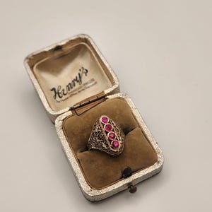 Può includere: Un anello color oro con quattro pietre rotonde di colore rosa vivace disposte in fila verticale su una fascia in metallo testurizzata e decorata. L'anello è esposto all'interno di un portagioie rettangolare aperto con interno in velluto beige e il testo Henry's.
