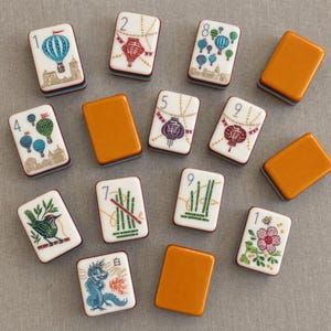 Puede incluir: Una colecci&oacute;n de fichas de mahjong rectangulares con caras blancas que muestran ilustraciones coloridas de globos aerost&aacute;ticos, linternas, bamb&uacute;, un p&aacute;jaro, una flor y un drag&oacute;n, junto a fichas con reverso naranja y los n&uacute;meros 1, 2, 4, 5, 7, 8 y 9.