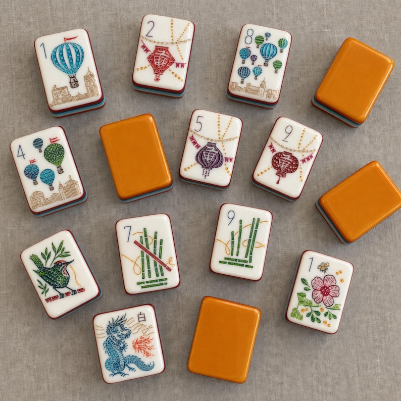 Puede incluir: Una colecci&oacute;n de fichas de mahjong rectangulares con caras blancas que muestran ilustraciones coloridas de globos aerost&aacute;ticos, linternas, bamb&uacute;, un p&aacute;jaro, una flor y un drag&oacute;n, junto a fichas con reverso naranja y los n&uacute;meros 1, 2, 4, 5, 7, 8 y 9.