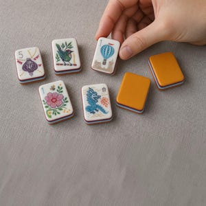 Puede incluir: Un conjunto de fichas de mahjong rectangulares con esquinas redondeadas, con ilustraciones coloridas de una linterna morada, un p&aacute;jaro verde, un globo aerost&aacute;tico azul, una flor rosa y un drag&oacute;n azul, junto a dos fichas de color amarillo mostaza.