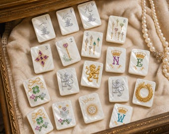 Può includere: Una collezione di tessere rettangolari in madreperla bianca con intricati disegni in metallo e pietre preziose, tra cui numeri, leoni, corone, motivi floreali e una tessera Joker, disposte su una superficie di tessuto morbido.