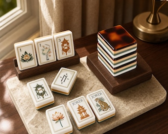 Puede incluir: Un juego de fichas de Mahjong rectangulares con ilustraciones detalladas de flores de loto, grullas, pagodas, tigres y la palabra JOKER. Las fichas blancas con detalles en color se exhiben sobre una superficie de piedra con soportes de madera.
