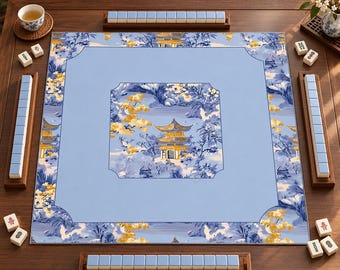 Può includere: Un tappetino da mahjong quadrato con un centro azzurro e un bordo decorato con un motivo paesaggistico blu e giallo raffigurante pagode e alberi. Quattro supporti in legno contenenti tessere azzurre sono posizionati attorno al tappetino.