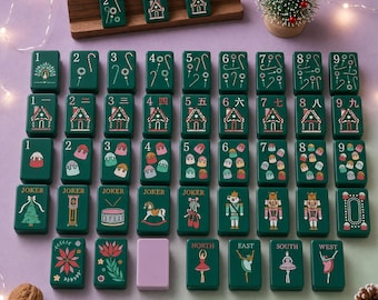Può includere: Un set di tessere da mahjong verde scuro con illustrazioni a tema natalizio, tra cui casette di pan di zenzero, bastoncini di zucchero, schiaccianoci e ballerine. Le tessere sono disposte in file, alcune su un supporto in legno.