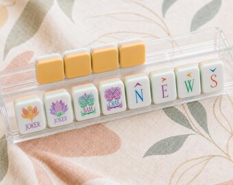 Può includere: Un supporto in acrilico trasparente contenente dodici tessere rettangolari da mahjong. La fila anteriore mostra tessere con illustrazioni colorate di fiori e creature, la parola JOKER e le lettere N, E, W, S. La fila posteriore ha quattro tessere gialle.